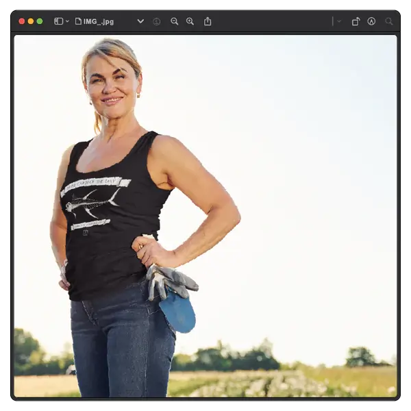 A person standing outdoors with one hand on their hip, wearing a black tank top and jeans. They hold gardening gloves, with a sunny sky and greenery in the background.