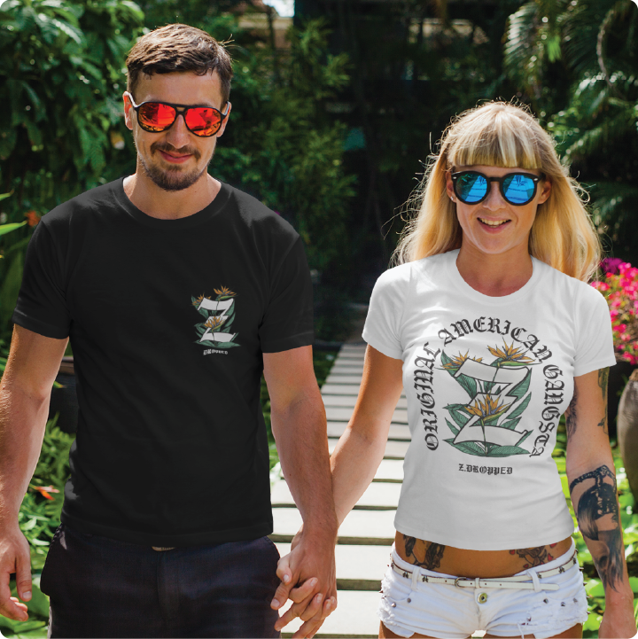 A smiling couple wearing sunglasses and matching graphic tees holding hands while walking on a path surrounded by greenery.