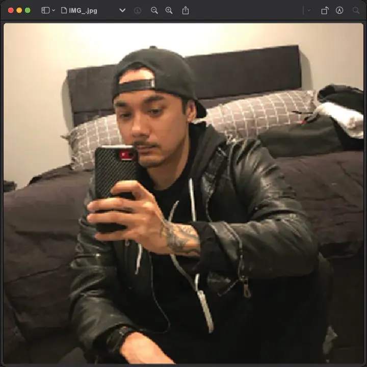 A person wearing a black leather jacket and cap takes a selfie in a bedroom. The phone case is black with a red accent.