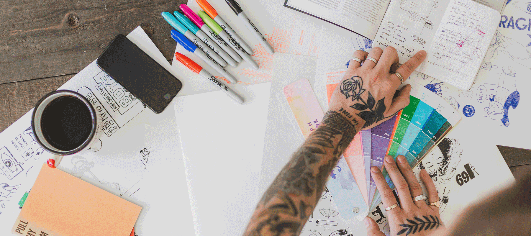 A person with tattooed arms working at a cluttered desk with a color swatch, markers, a cup of coffee, and various papers and notebooks.