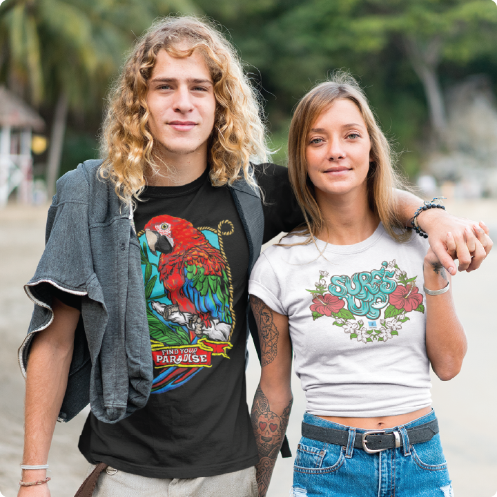 A man and woman smiling on a beach. The man has long curly hair and wears a black t-shirt with a parrot design. The woman has straight hair and wears a white t-shirt with the word Surf.