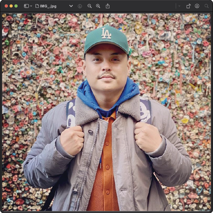 A person in a green cap and layered clothing stands in front of a colorful gum wall, holding the straps of their backpack.